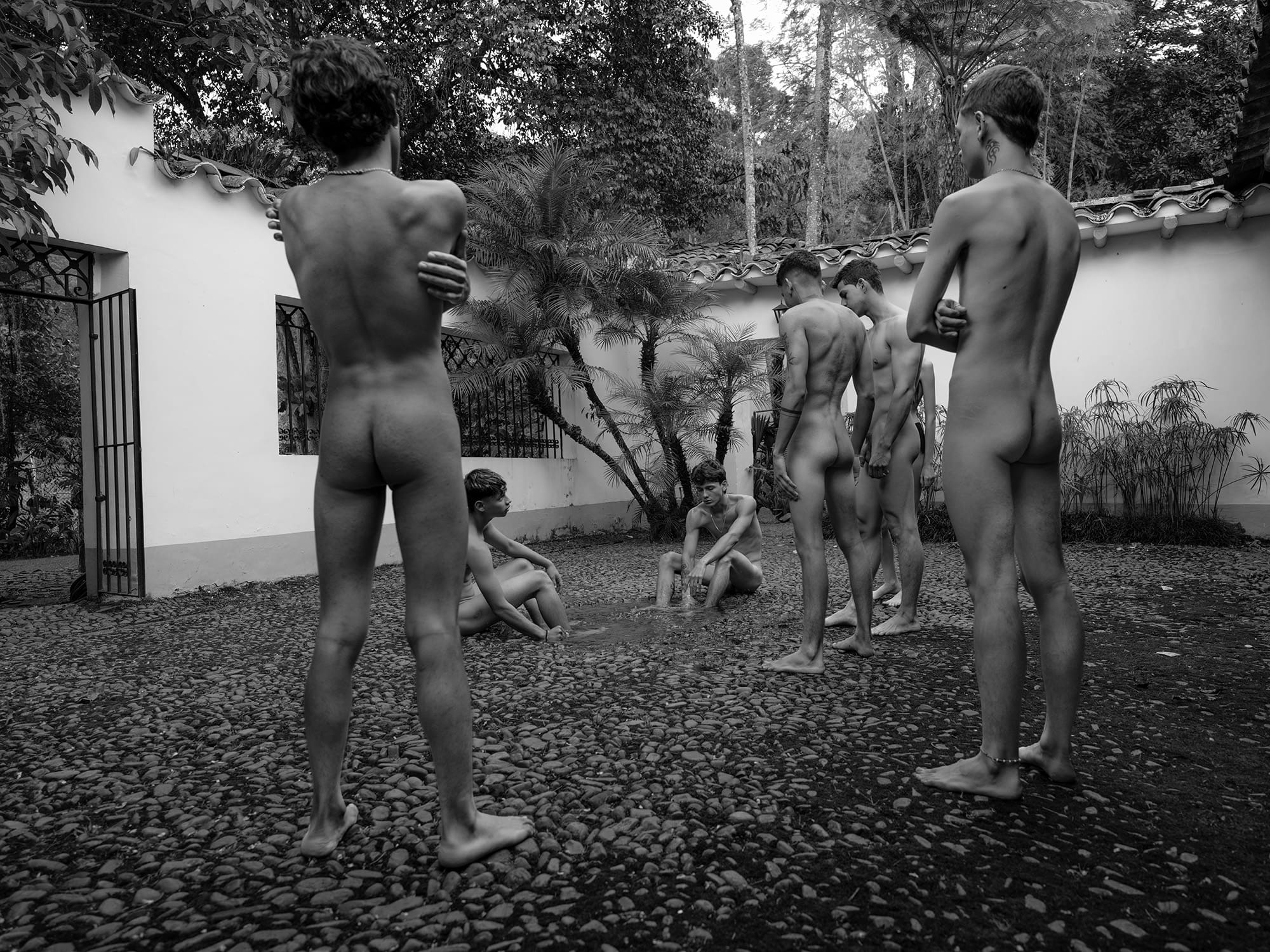 Black and white photograph of six young men in swimwear gathered in the stone-paved courtyard of a Colombian finca, surrounded by whitewashed walls and tropical vegetation. Some stand, some crouch or sit. Their backs are mostly turned to the camera. The atmosphere is tense and unresolved.